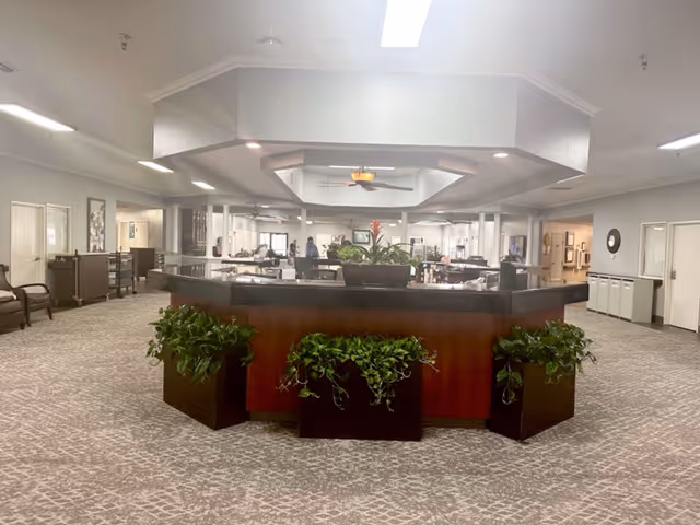 Interior view of a senior living facility reception area with a large octagonal wooden desk in the center, decorated with green plants. The space is well-lit with ceiling lights and a skylight, featuring carpeted floors and seating areas along the walls.