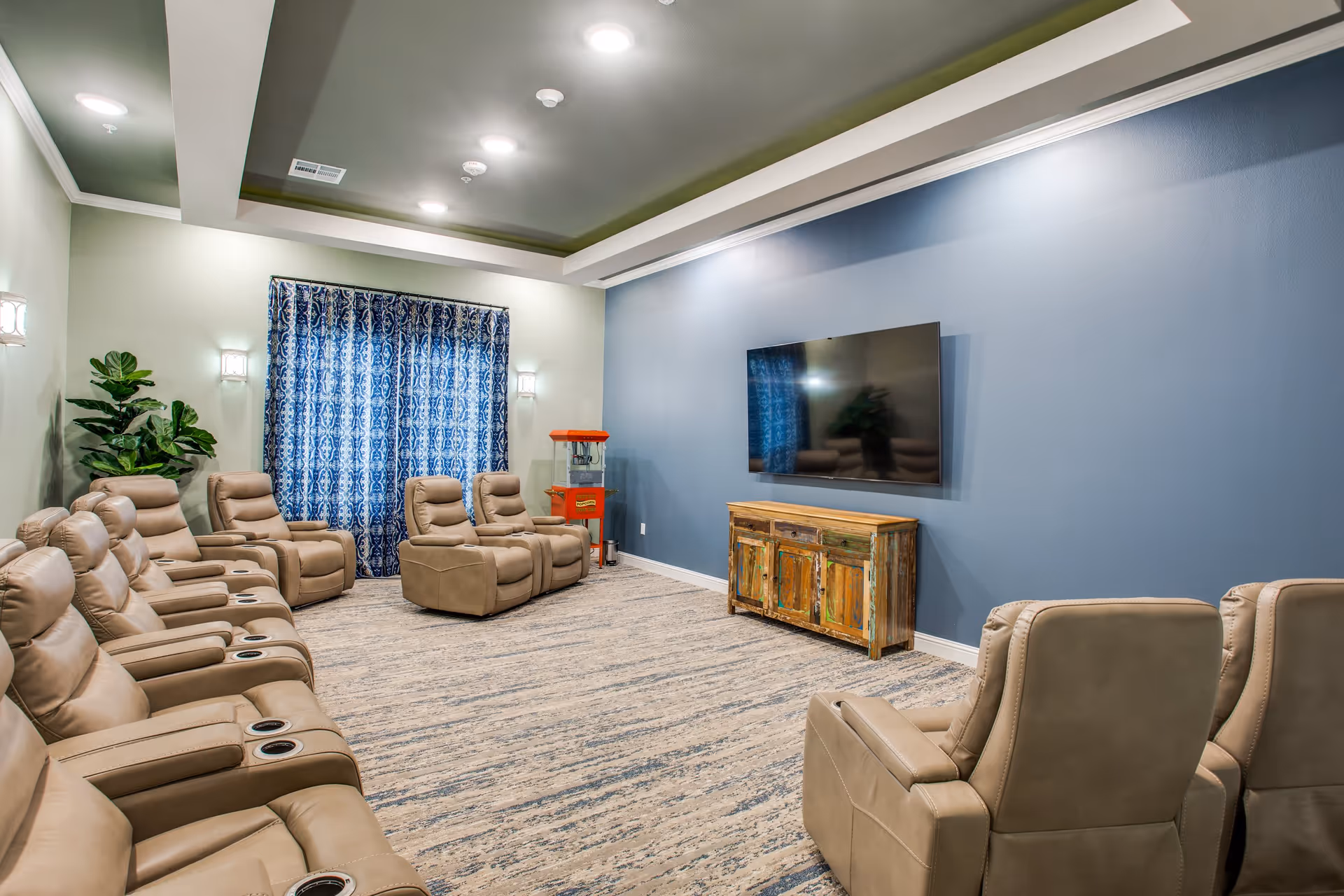 A cozy senior living facility lounge with beige recliner chairs arranged in rows facing a large flat-screen TV mounted on a blue accent wall. The room has a patterned carpet, blue and white patterned curtains, a small popcorn machine in the corner, and a rustic wooden cabinet beneath the TV. There is a green potted plant near the window and soft ceiling lights illuminating the space.