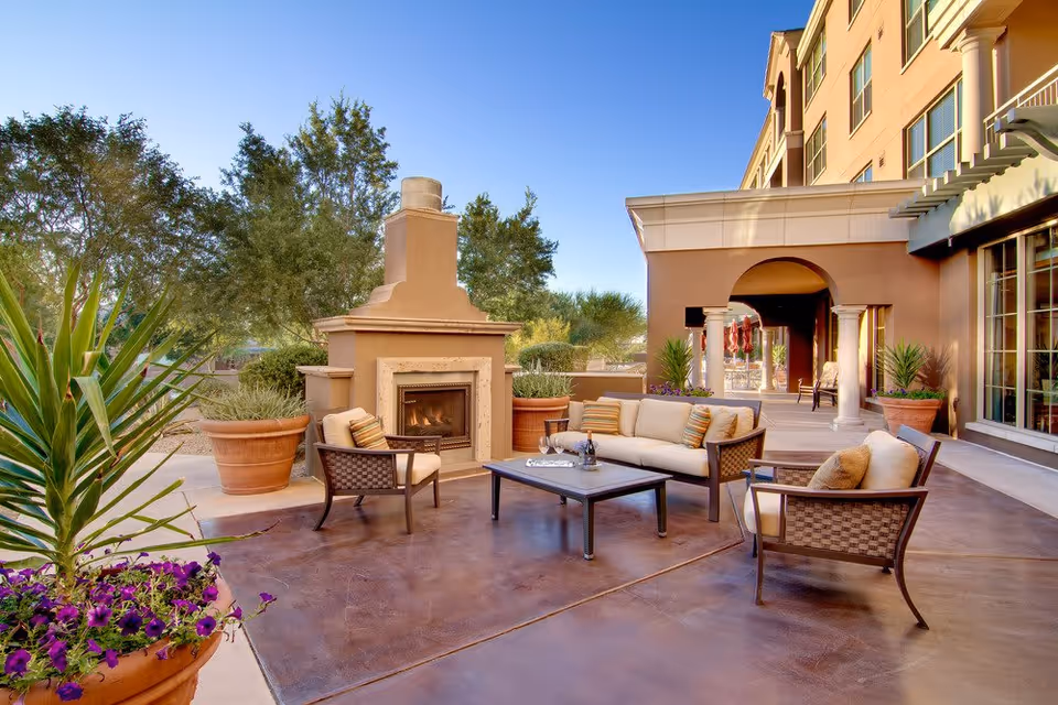 Outdoor patio area with cushioned seating including a sofa and two armchairs arranged around a coffee table. There is a large outdoor fireplace in the background, surrounded by potted plants and trees. The patio is adjacent to a building with large windows and an arched covered walkway.