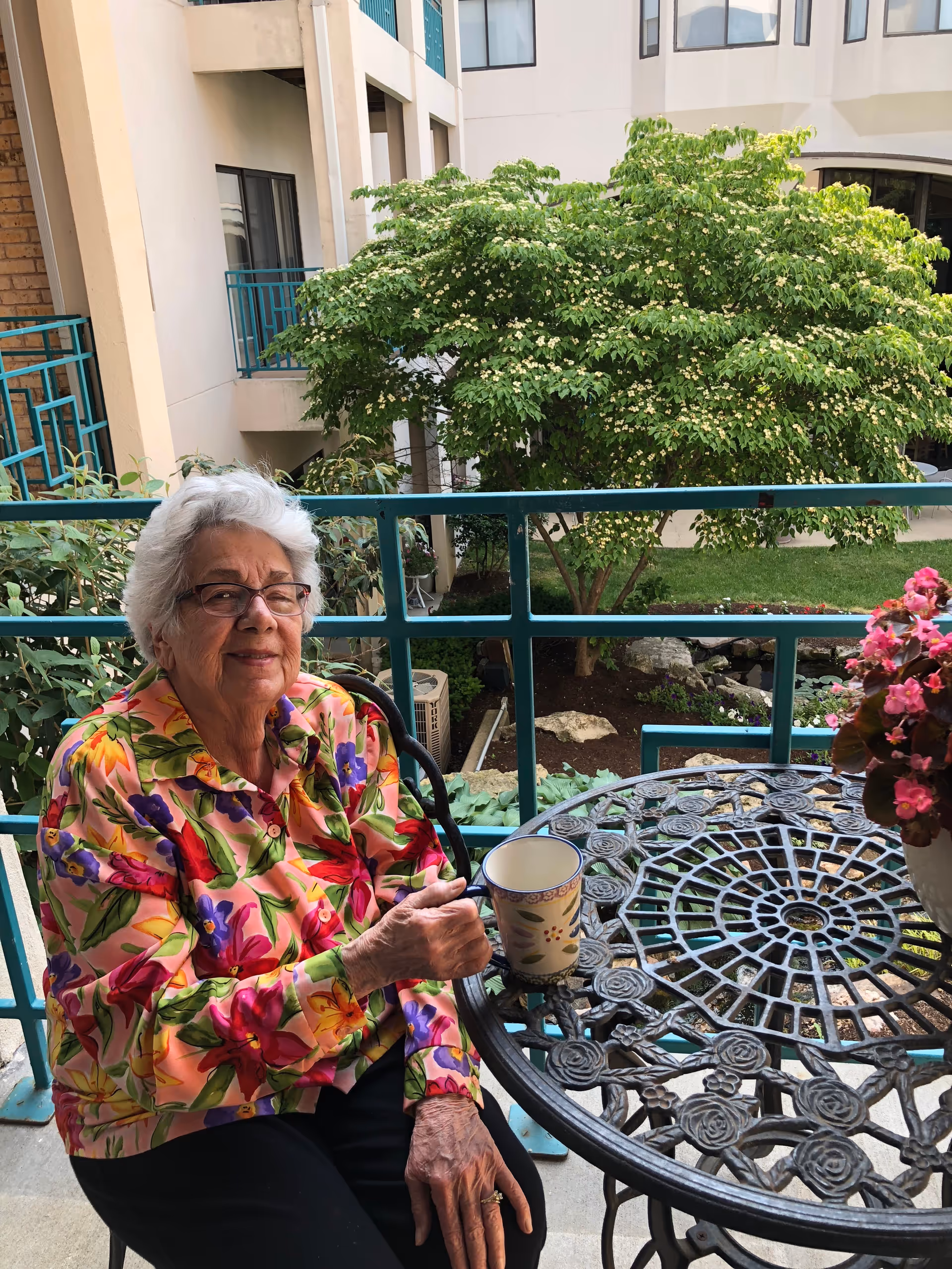An elderly woman with white hair and glasses sits on a chair on a balcony. She is wearing a colorful floral jacket and holding a cup. The balcony has a decorative metal table with a potted pink flower and green railing. Behind her, there is a view of a courtyard with green trees and a building with windows and balconies.