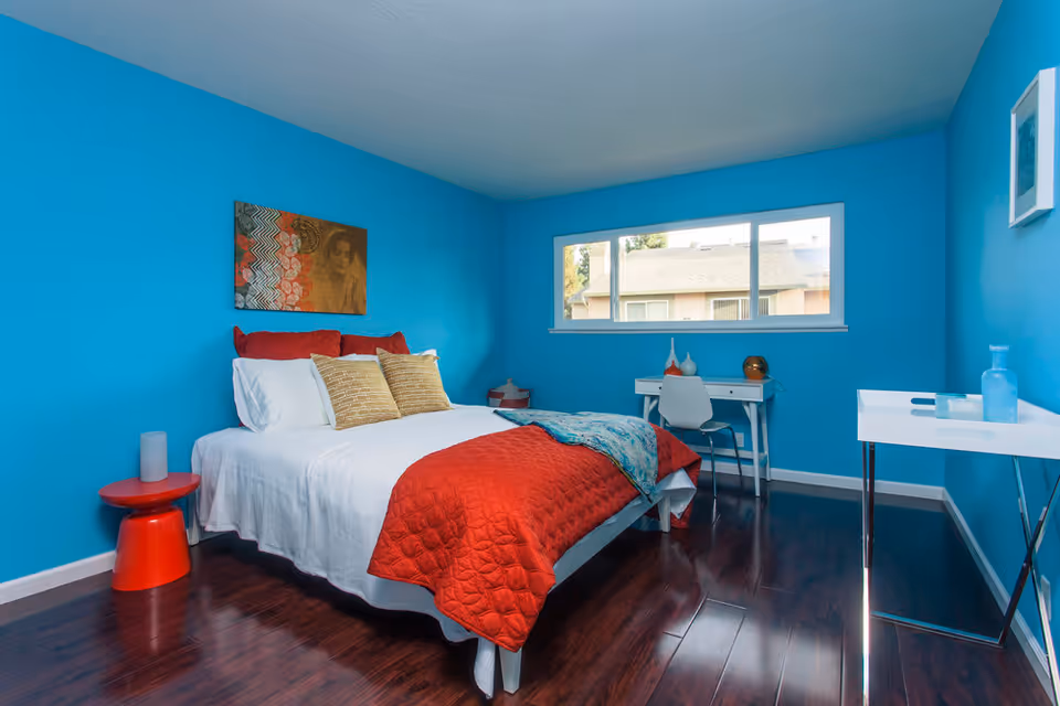 Bright turquoise bedroom with a bed dressed in white linens and a red quilt, a small desk under a window, and dark hardwood floors.