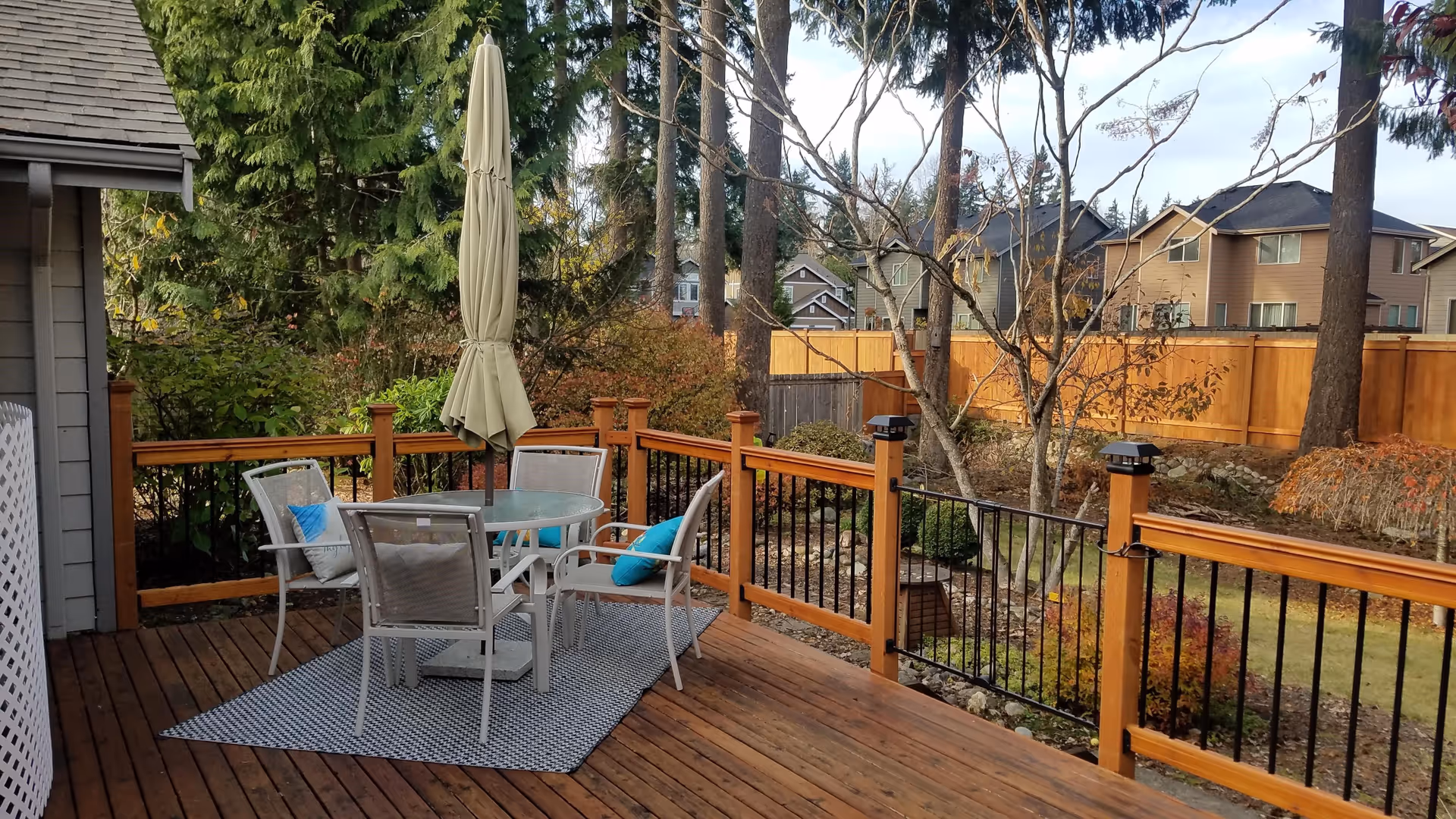 A wooden deck with a round glass table and four chairs, each with a cushion. A closed beige patio umbrella stands in the center of the table. The deck is surrounded by a wooden railing with black metal balusters. In the background, there are trees, bushes, and a wooden fence with houses visible beyond it.