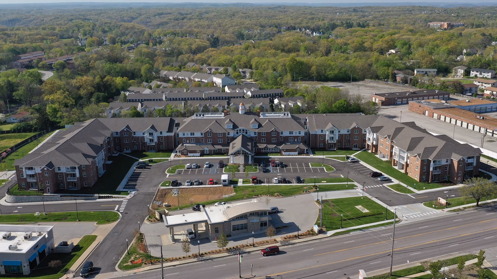 Aerial view of Creek Valley Retirement Community showing a large, multi-wing brick building with multiple parking lots and surrounding roads. The building is surrounded by green lawns and trees, with a smaller building in front near the main road. Residential houses and wooded areas are visible in the background.
