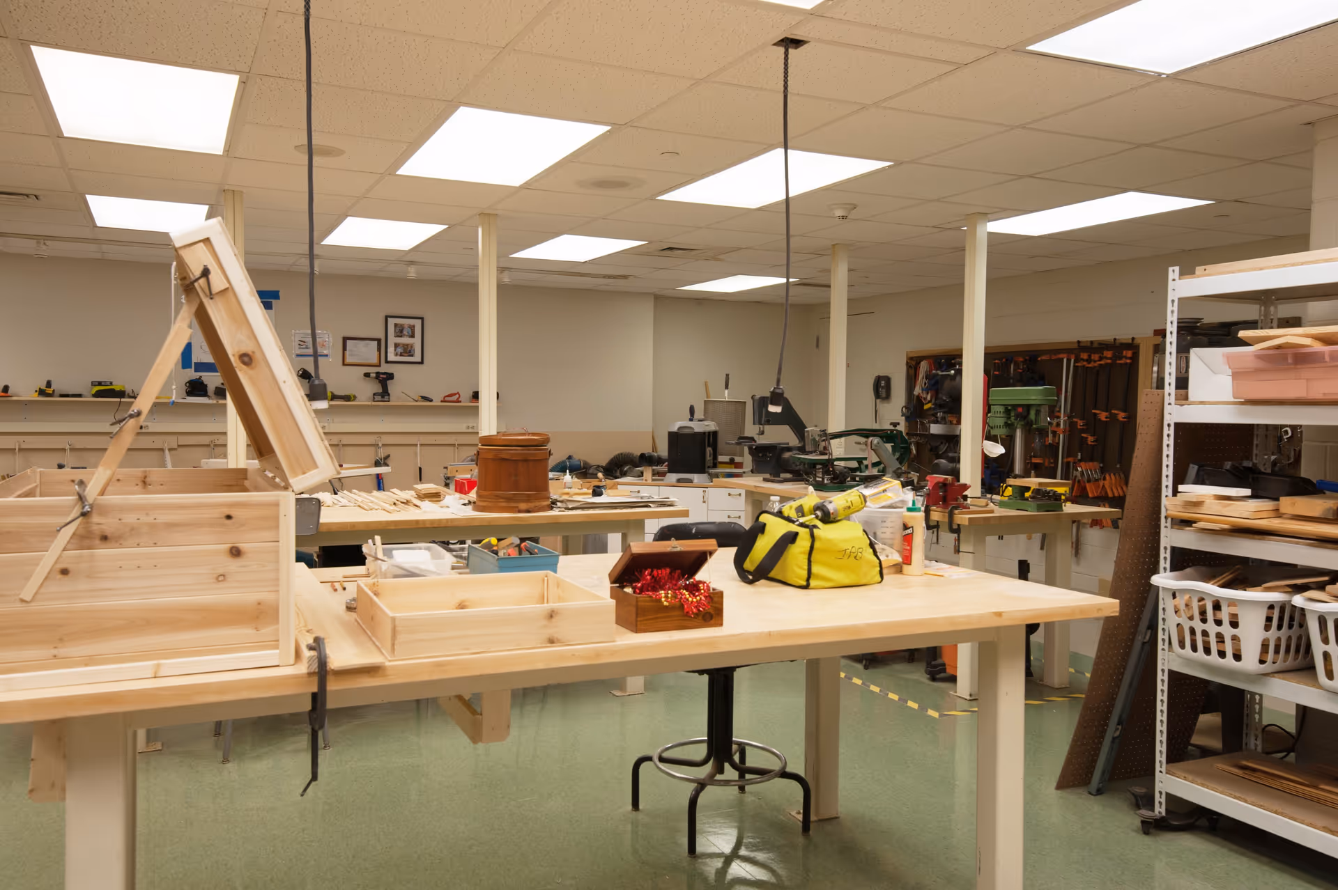 Interior view of a woodworking workshop with wooden tables, various woodworking tools, clamps, and shelves holding supplies. The room has fluorescent ceiling lights and a green floor.