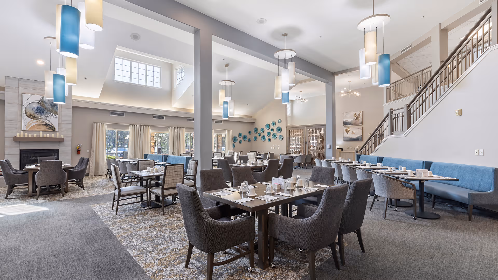A spacious, well-lit dining room with multiple tables and chairs arranged neatly. The room features high ceilings with modern pendant lights in blue and white, large windows with curtains allowing natural light, a fireplace with artwork above it, and a staircase with a railing on the right side. The tables are set with cups, plates, and napkins, ready for dining.