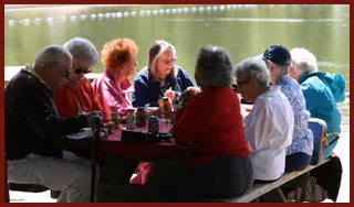 Elderly residents gathered around an outdoor picnic table by a lake, sharing drinks and conversation.