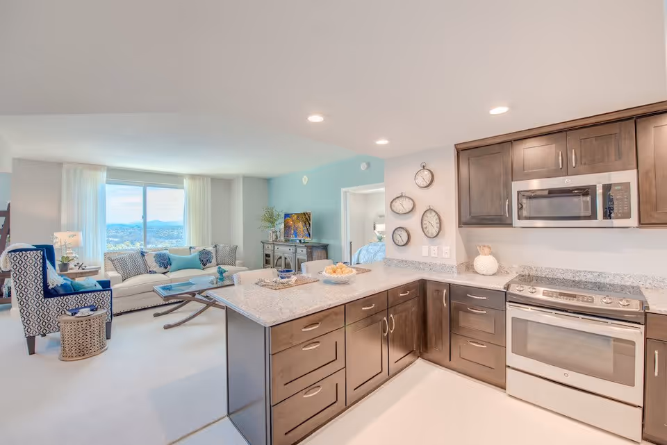 Bright and modern open-concept living space featuring a kitchen with dark wood cabinets, granite countertops, stainless steel microwave and oven. The kitchen island has a bowl of fruit and decorative items. Adjacent living room has a large window with a scenic view, a white sofa with patterned pillows, a blue patterned armchair, a coffee table, and a TV on a wooden console. A bedroom is partially visible through an open door.