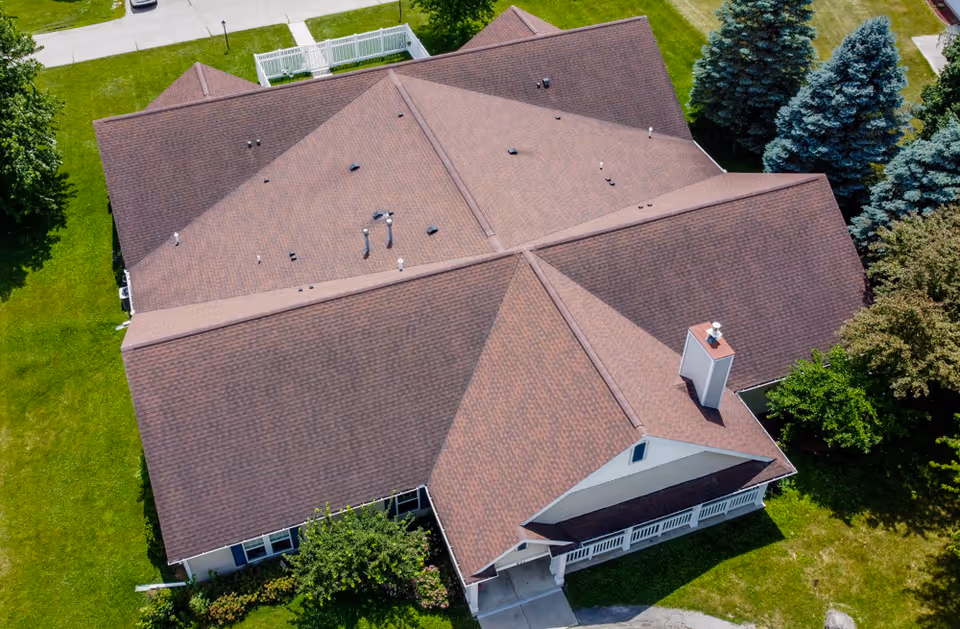 Aerial view of a large brown-shingled building roof with surrounding green lawn and trees.