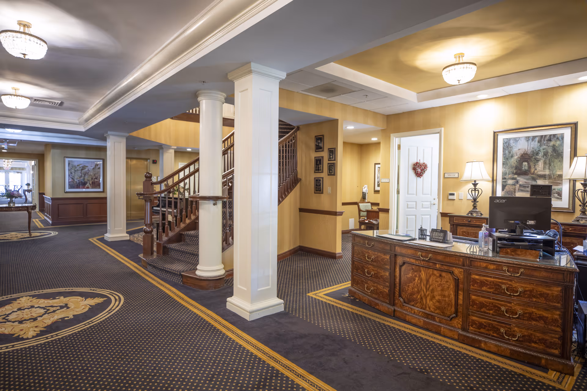 Interior view of a senior living facility lobby with a wooden reception desk, two table lamps, a computer, and a staircase with wooden railings. The walls are painted yellow with framed artwork and plaques. The carpet is dark with a patterned design and there are white columns supporting the ceiling with chandeliers.