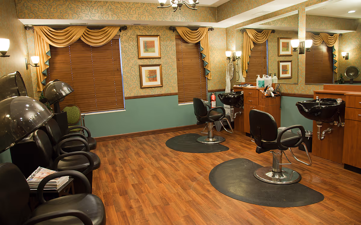 Interior view of a senior living facility's hair salon with wooden flooring, two black salon chairs in front of sinks, multiple black hair dryer chairs along the left wall, decorative curtains on windows, framed artwork on the walls, and a large mirror on the right side.