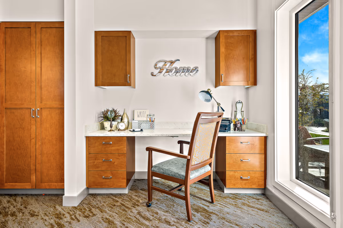 Small built-in wooden desk and cabinets with a chair, lamp and decorative "Home" sign next to a large window.