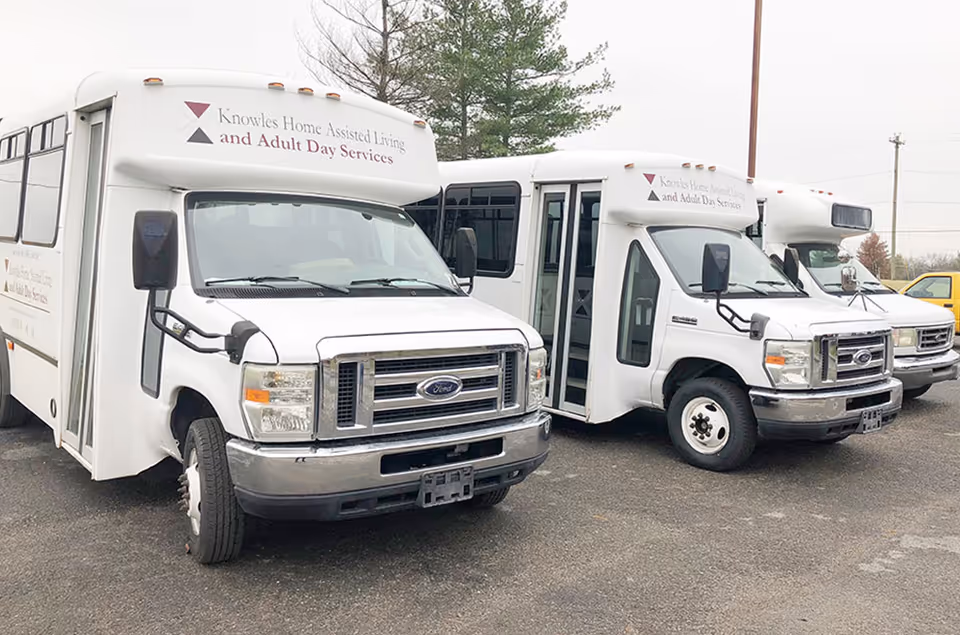 Three white shuttle buses parked side by side in a parking lot. The buses have signage on the front indicating they belong to Knowles Home Assisted Living and Adult Day Services. Trees and a cloudy sky are visible in the background.