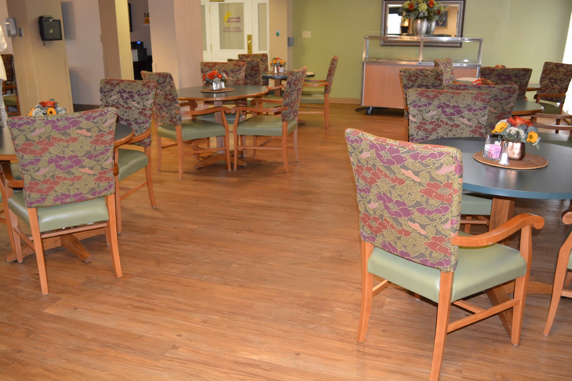 Dining room with round tables, patterned upholstered chairs, floral centerpieces, and wood flooring.