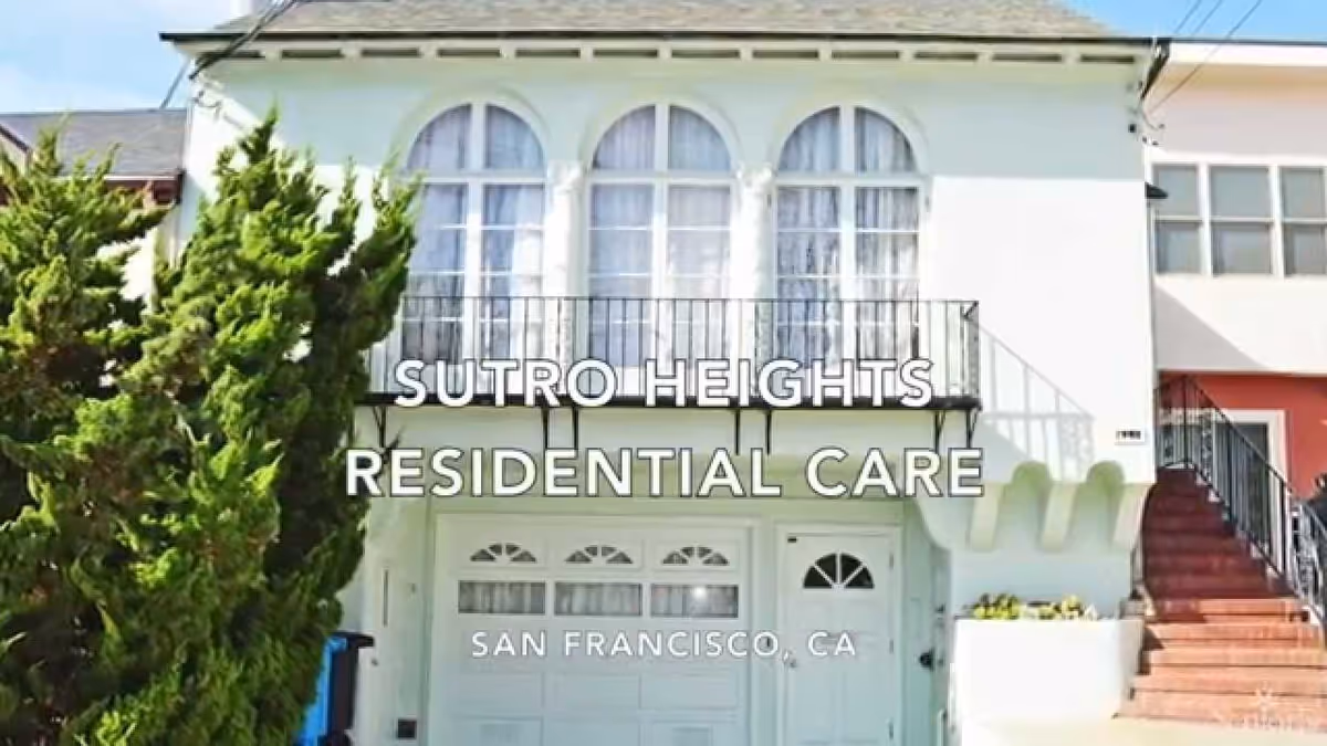 Front exterior view of a white residential building with three large arched windows on the upper floor, a balcony with a black railing, a garage door, and an entrance door below. There are green trees on the left side and stairs leading up on the right side. Text overlay reads 'Sutro Heights Residential Care San Francisco, CA'.