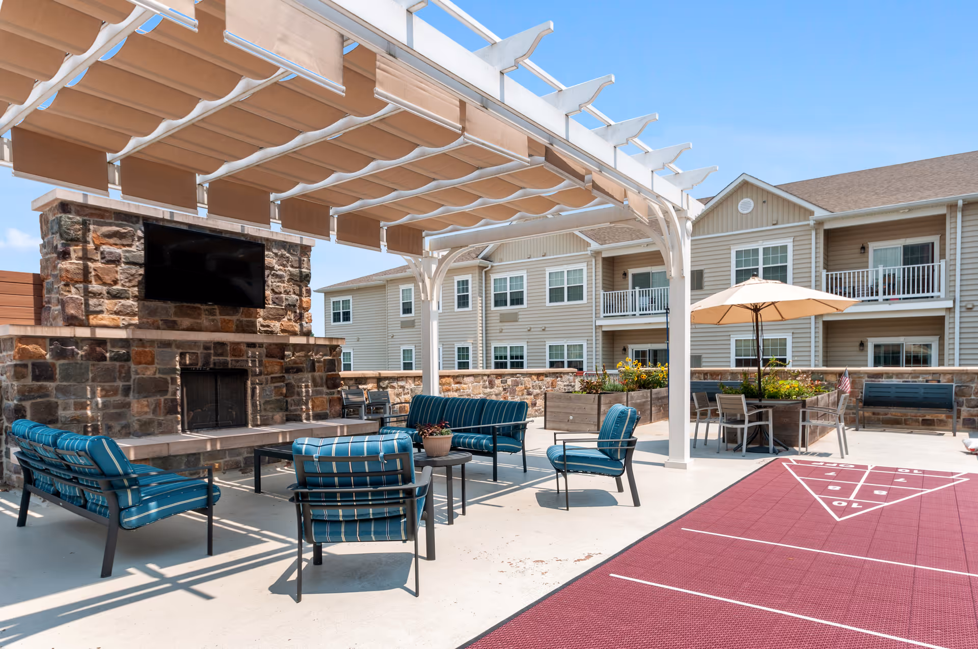 Outdoor patio area at Merrill Gardens at West Chester featuring a stone fireplace with a mounted TV, blue cushioned seating under a white pergola with retractable fabric shades, a shuffleboard court, and a table with an umbrella. The background shows a two-story building with balconies and windows.