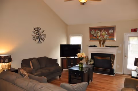 A cozy living room with two brown sofas, a black TV stand with a flat-screen TV, a black coffee table with a flower vase, a fireplace with floral artwork above it, and a door with an exit sign. The room has wooden flooring and beige walls.