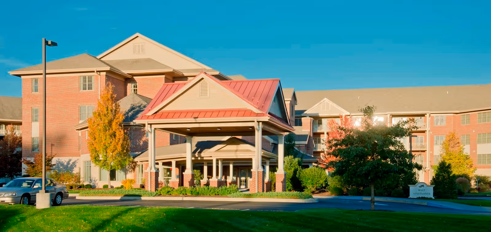 Exterior view of a senior living facility named Bethany Village West, showing a large brick building with multiple floors, a covered entrance with a red roof, landscaped greenery, trees with autumn foliage, a parked car, and a clear blue sky.