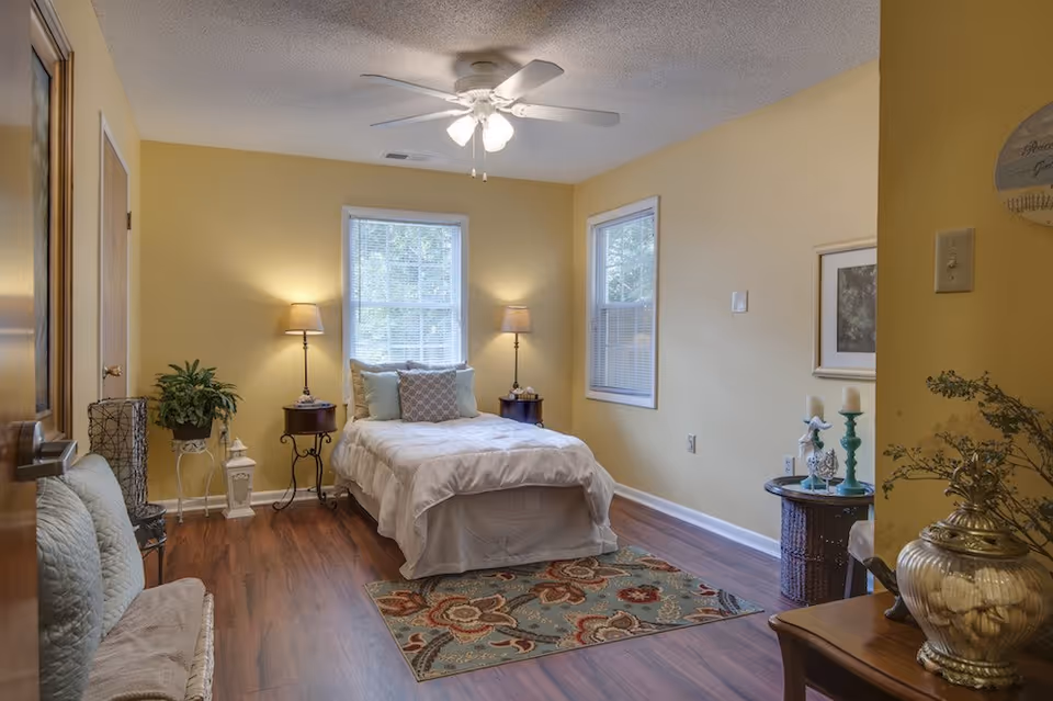 A cozy bedroom with yellow walls and wooden flooring. The room features a bed with white bedding and multiple pillows, positioned between two windows with blinds. There are two bedside tables, each with a lamp. A colorful patterned rug is placed at the foot of the bed. Additional furniture includes a cushioned bench, a small round table with decorative items, and a plant stand with a green plant. The ceiling has a white fan with lights.
