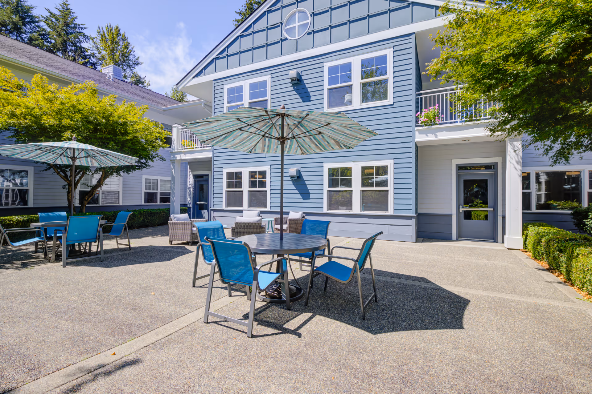 Sunny outdoor courtyard with blue patio tables, umbrellas, and chairs in front of a two-story blue-sided assisted living building.