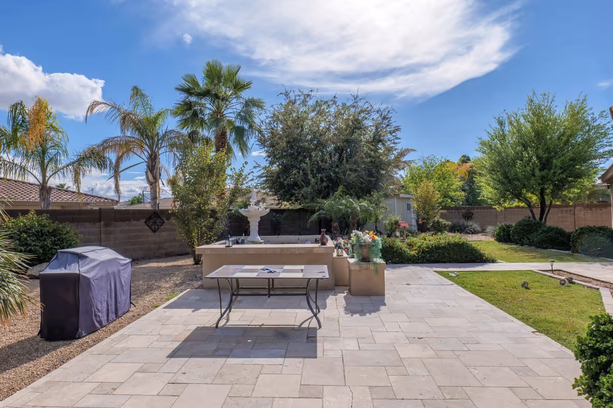 Backyard patio with a table and covered grill, a fountain, palm trees, and landscaped garden under a blue sky.