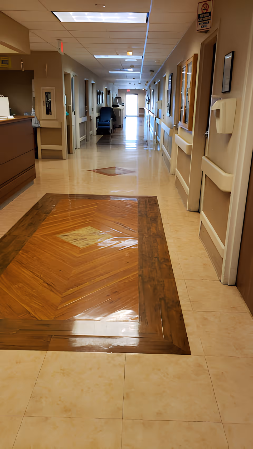 A long, clean hallway in a healthcare or rehabilitation facility with beige walls and a polished floor featuring a wooden inlay design. The hallway has several doors on both sides, handrails along the walls, ceiling lights, and a chair positioned near the far end. There is a reception desk on the left side near the foreground.
