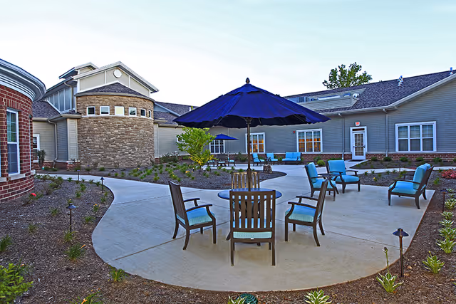 Outdoor courtyard area of a senior living facility with a circular concrete patio featuring several chairs with blue cushions and a table with a large blue umbrella. Surrounding the patio are landscaped garden beds and the exterior of the facility buildings with windows and doors.