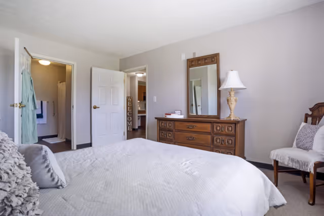 A bedroom with a large bed covered in a white quilt and decorative pillows. There is a wooden dresser with a mirror and a lamp on top, and a chair with a cushion and throw blanket to the right. Two doors are visible, one leading to a bathroom with a shower curtain and towels, and the other leading to another room with a 'WELCOME' sign on the door frame.