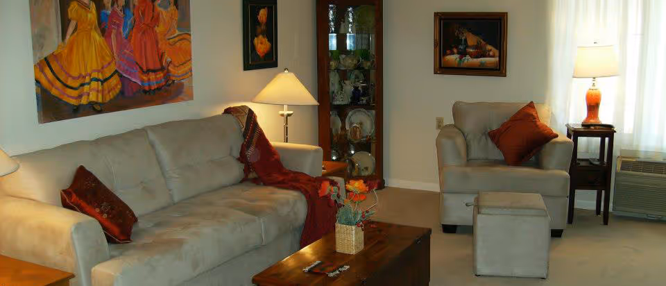 A cozy living room with a beige sofa and armchair, both adorned with red throw pillows. A red throw blanket is draped over the sofa. There is a wooden coffee table with a small decorative basket and flowers. Two lamps with beige and orange bases provide warm lighting. A glass cabinet displaying dishes is against the wall, and colorful paintings hang above the sofa and armchair. A window with white curtains and an air conditioning unit is visible on the right side.