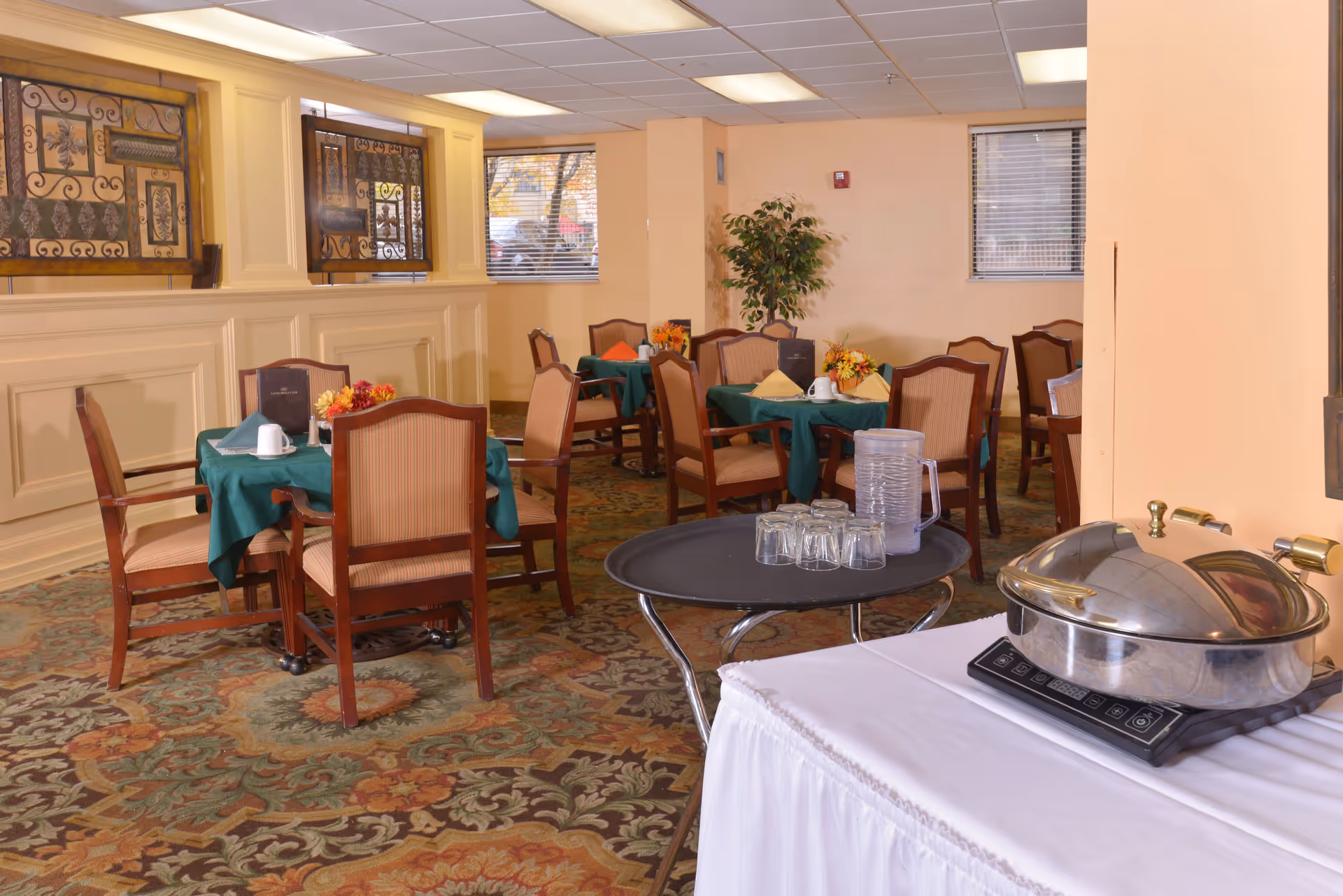 A dining room in a senior living facility with several tables covered in green tablecloths, each set with cups, napkins, and small floral centerpieces. There are wooden chairs with cushioned seats around the tables. A serving station with a covered chafing dish and a tray with a pitcher and glasses is visible in the foreground. The room has beige walls, windows with blinds, and decorative wall art.