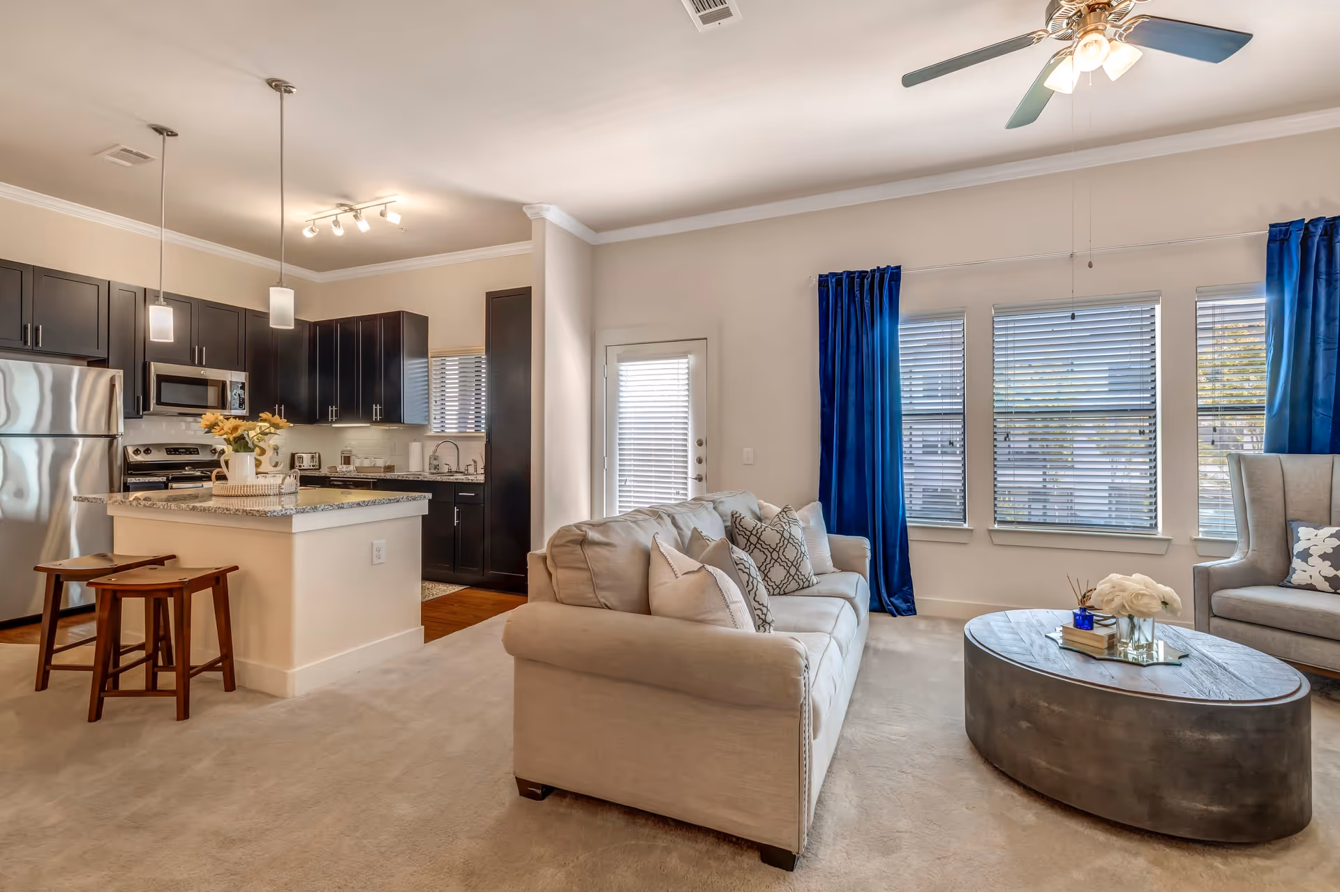 A bright and modern living room and kitchen area featuring a beige sofa with patterned pillows, a round coffee table with decorative items, and a gray armchair. The kitchen has dark cabinets, stainless steel appliances, a granite countertop island with two wooden stools, and pendant lighting. Large windows with blue curtains and blinds allow natural light to fill the space, and a ceiling fan with lights is mounted on the ceiling.
