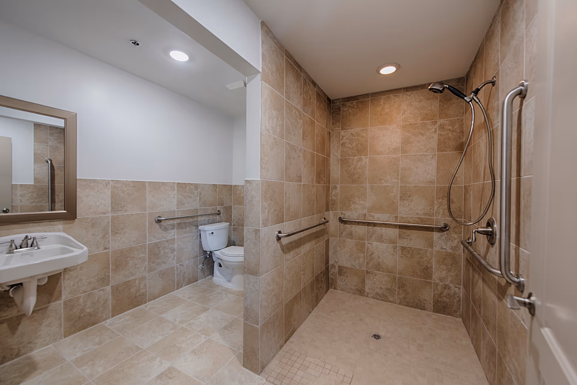 A spacious bathroom with beige tiled walls and floor, featuring a walk-in shower with a handheld showerhead and multiple grab bars, a white toilet with a grab bar beside it, and a white sink with a mirror above it. The bathroom is well-lit with recessed ceiling lights.
