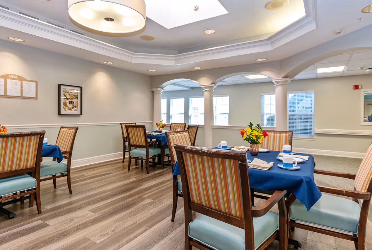 A bright dining room with several tables covered in blue tablecloths, each set with plates, cups, and napkins. The chairs have wooden frames with striped upholstery. The room features a skylight, recessed lighting, columns, and large windows letting in natural light. A menu board and framed pictures are visible on the wall.