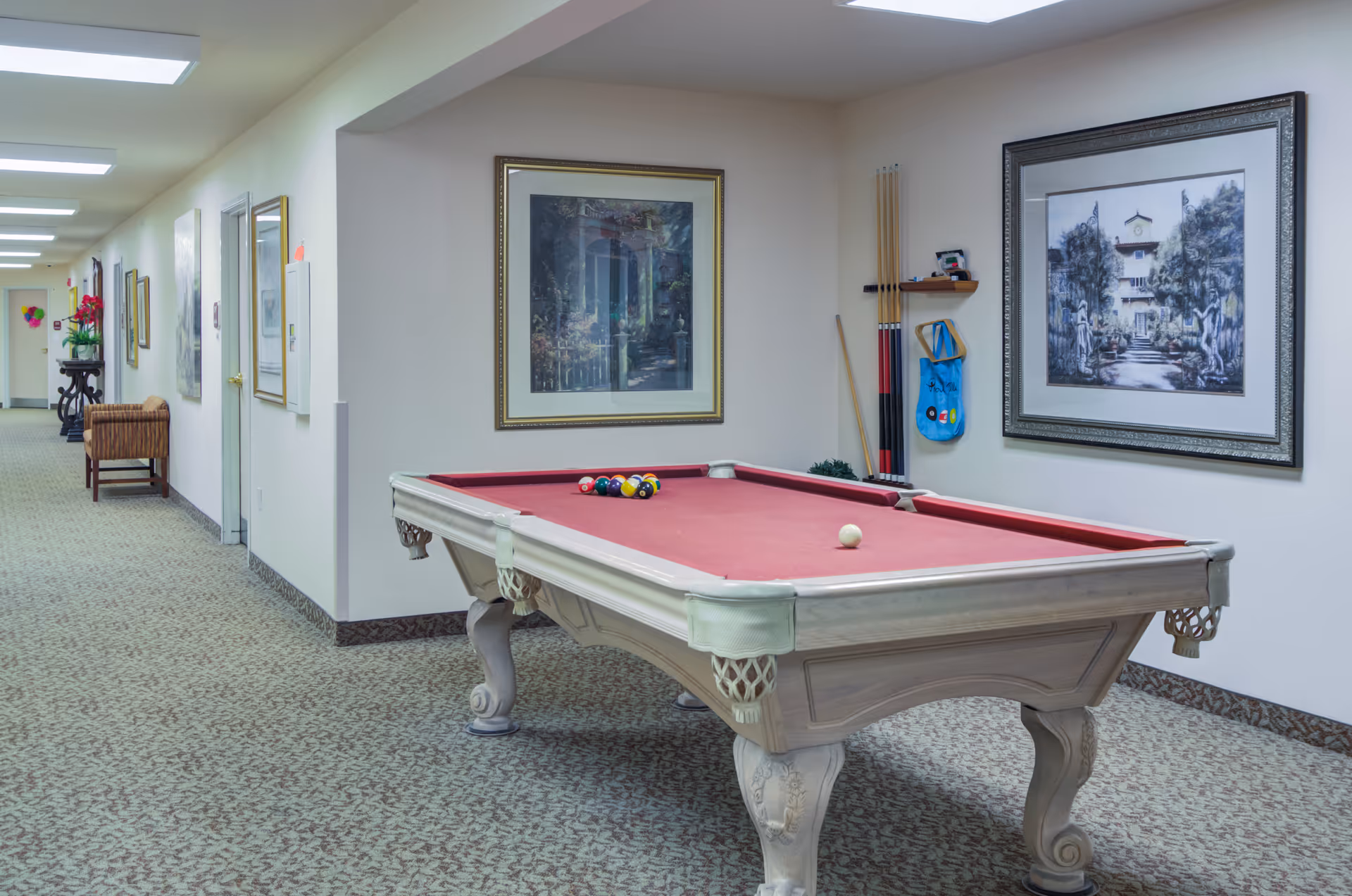 Interior hallway of a senior living facility featuring a pool table with a red felt surface. The pool table is set up with billiard balls and cues on a wall rack. The hallway has framed artwork on the walls, carpeted flooring, and overhead fluorescent lighting. There are chairs and decorative plants along the hallway.