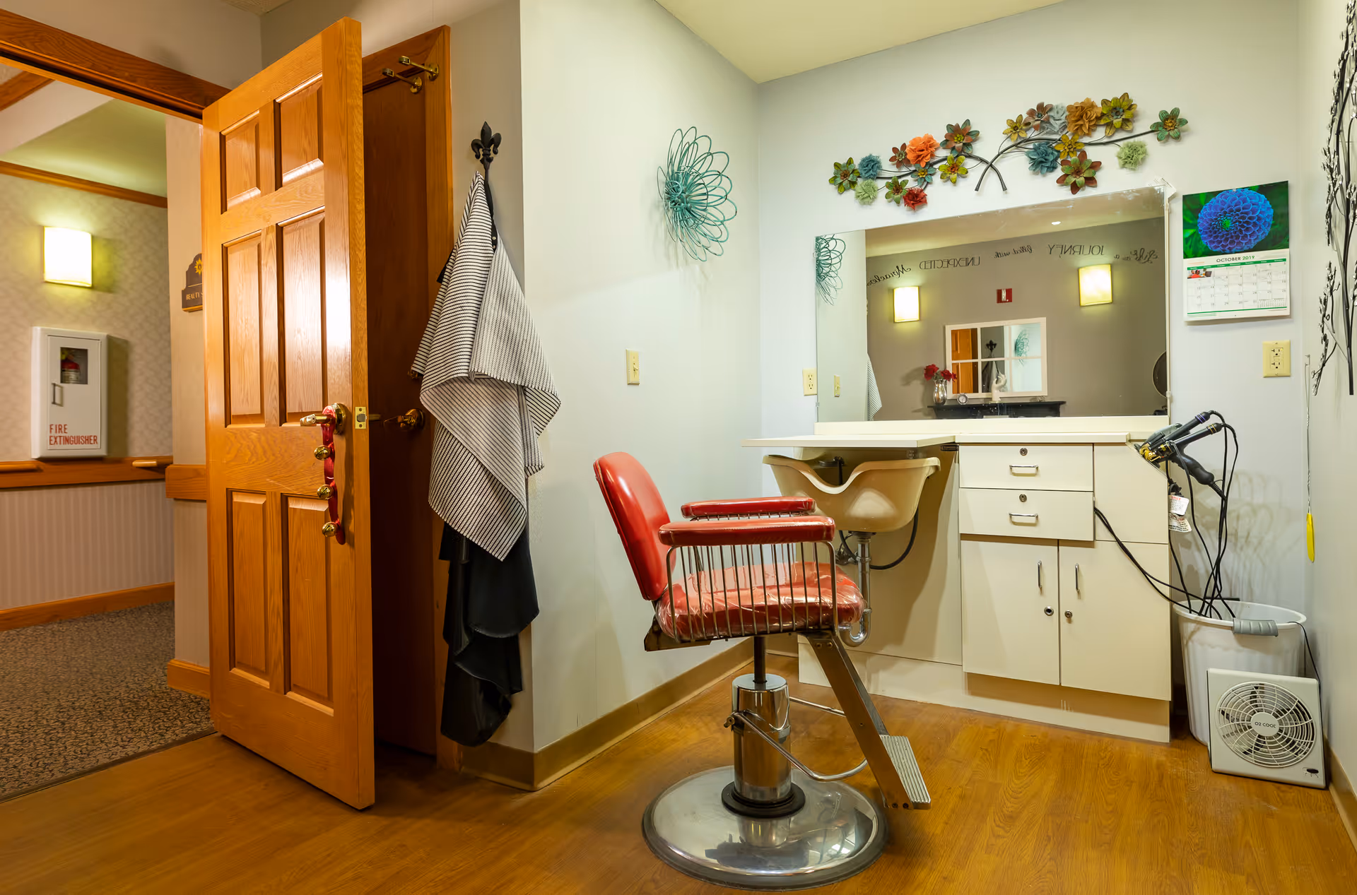 A small salon area inside Homestead Assisted Living of Kearney featuring a red salon chair in front of a white vanity with drawers and a large mirror. The wall above the mirror is decorated with colorful metal flower art. A hairdryer is placed on the right side of the vanity, and a calendar is hanging on the wall. The door to the room is open, showing a hallway with a fire extinguisher cabinet.