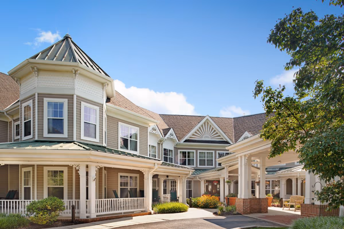 Exterior view of a senior living facility building with beige siding, white trim, and a covered entrance. The building features a turret with multiple windows and a porch with white railings. There are green bushes and trees around the entrance under a clear blue sky.