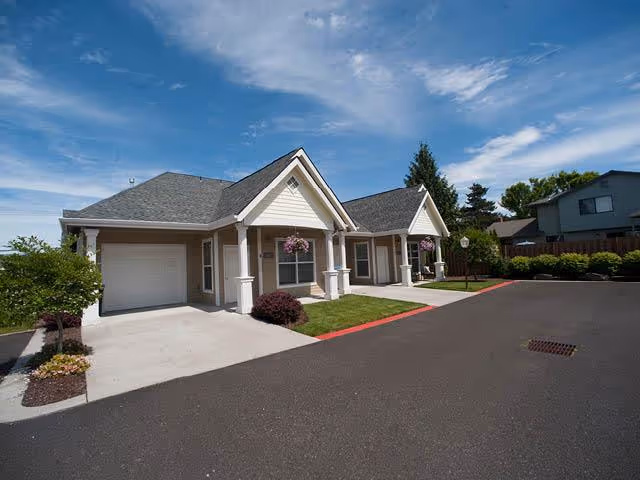 Exterior view of a single-story residential building with a garage, manicured lawn, hanging flower baskets, and a paved driveway under a blue sky with some clouds.