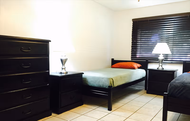 A clean bedroom with two single beds, dark wood dressers and nightstands topped with lamps and a window with blinds.