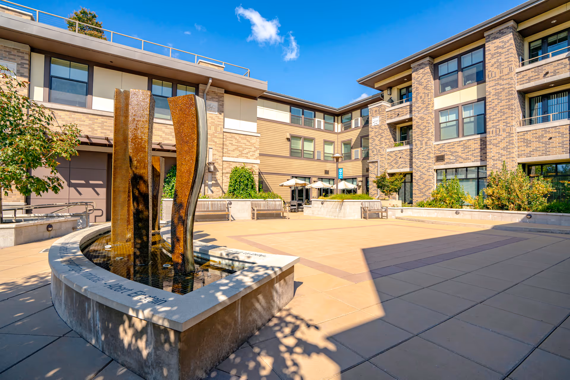 Outdoor courtyard area at Merrill Gardens at Lafayette featuring a modern water fountain with three vertical curved panels, surrounded by benches and landscaping. The courtyard is enclosed by a multi-story brick and siding building under a clear blue sky.