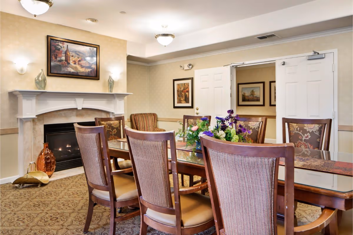 A cozy dining room with a glass-top wooden table surrounded by six upholstered chairs. A floral centerpiece decorates the table. The room features a fireplace with a white mantel, two decorative vases, and a framed painting above it. The walls have light-colored wallpaper with framed artwork, and double white doors are open to an adjacent room.
