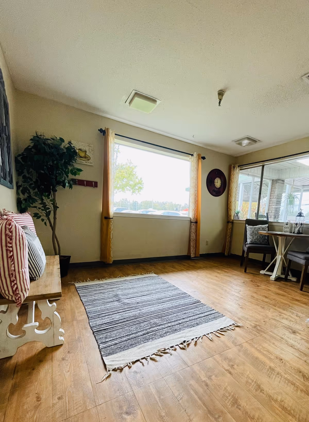 Bright communal sitting area with wooden floor, striped rug, a bench with pillows, a potted plant, and a small table with chairs by large windows.