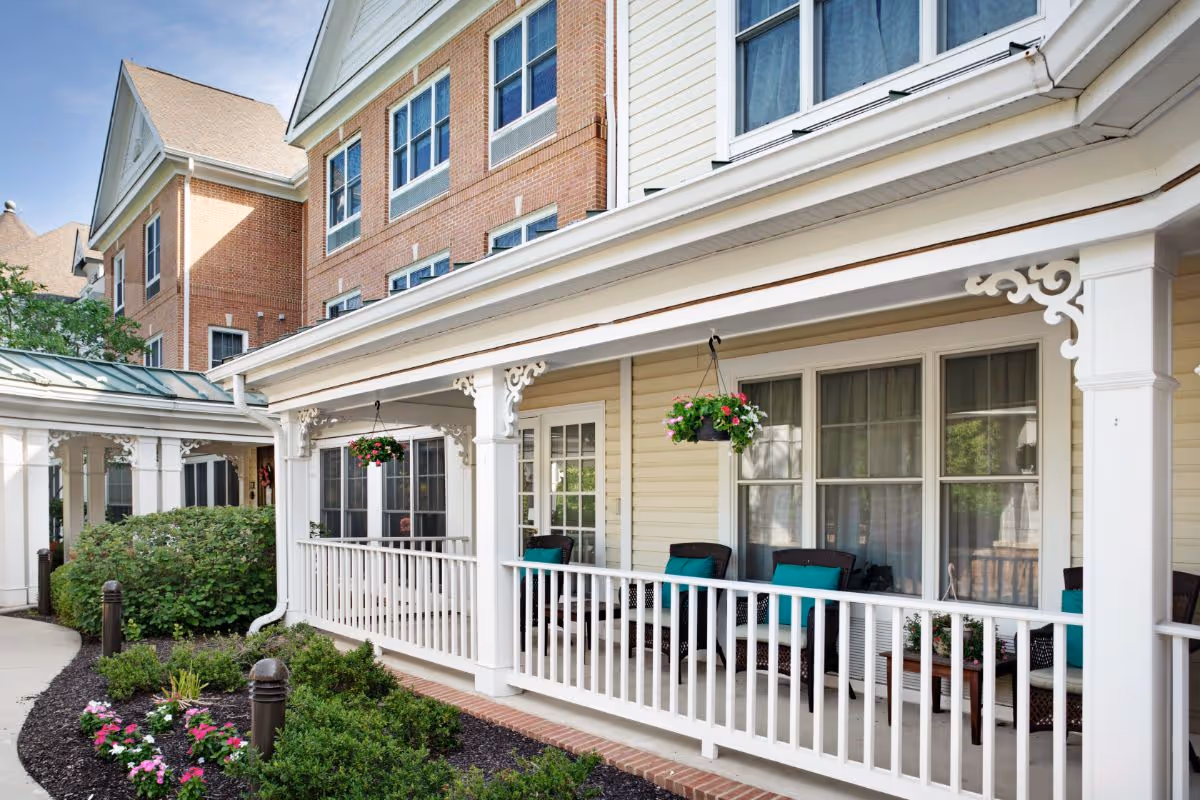 Exterior view of a senior living facility porch area with white railing, hanging flower baskets, and wicker chairs with teal cushions. The building has beige siding and large windows with curtains. There are well-maintained bushes and flowers along the walkway.