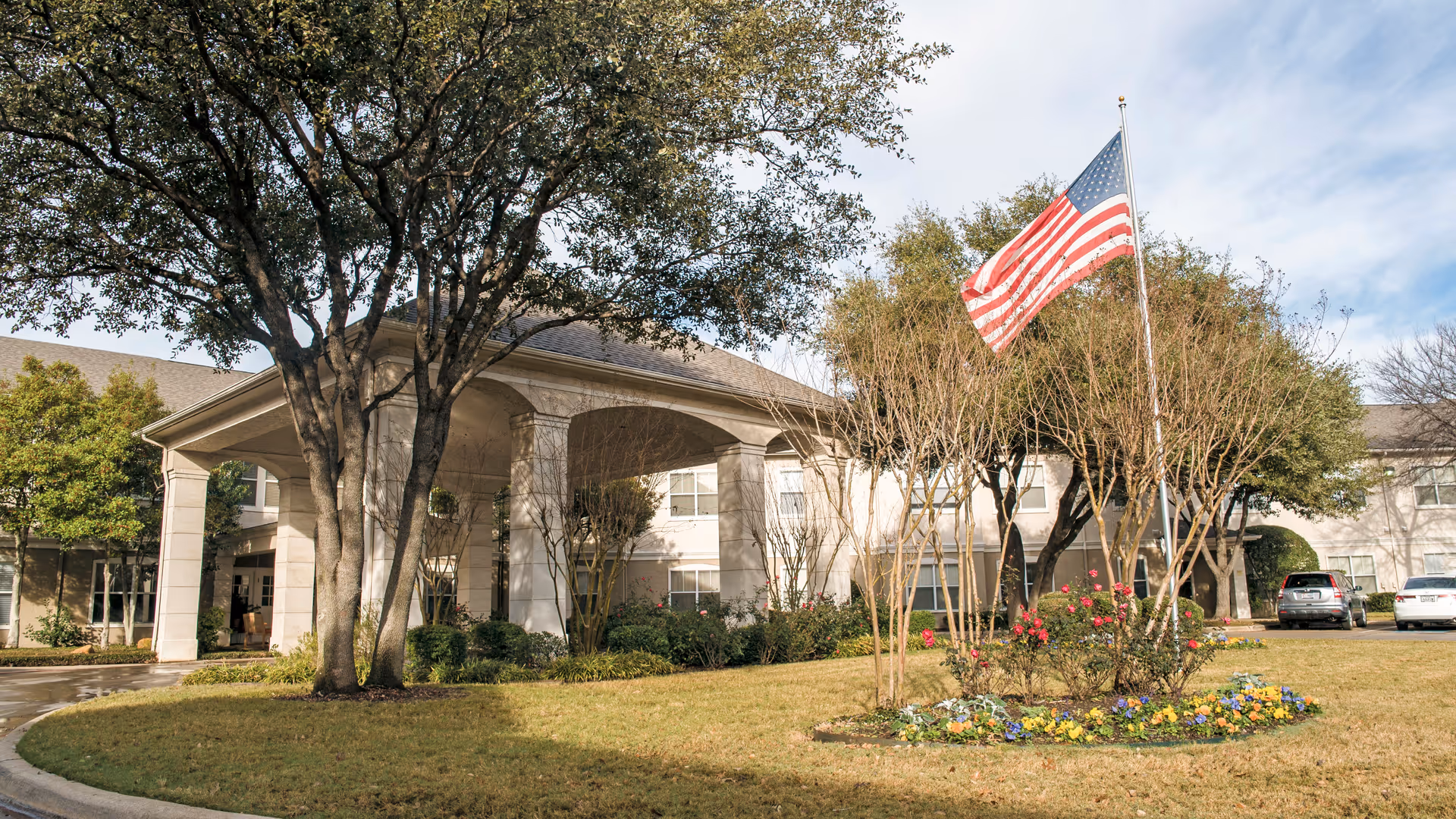 Exterior view of Brookdale Eden Estates senior living facility showing the entrance with a covered driveway, trees, a flagpole with an American flag, flower beds, and parked cars.