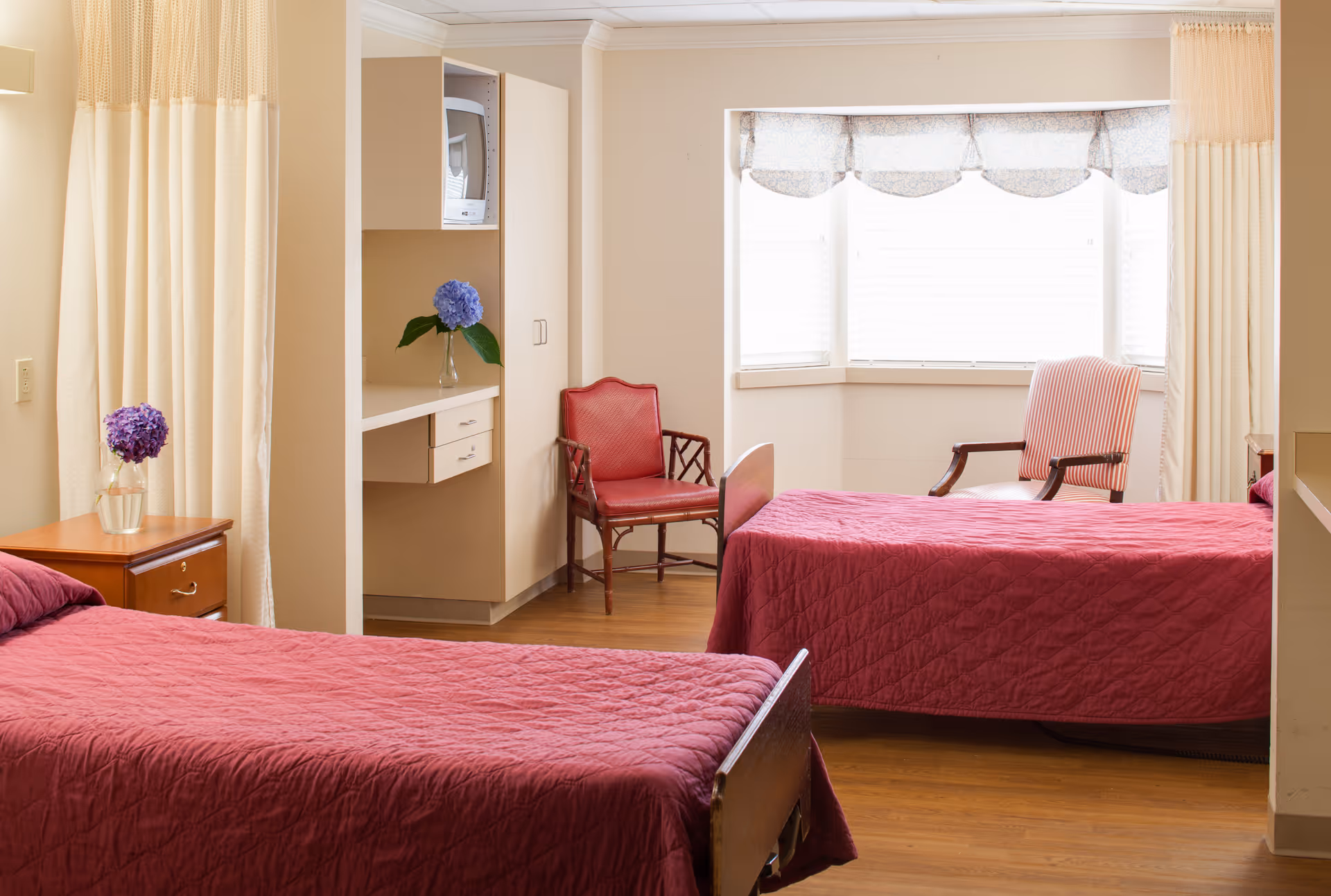 A bright room with two single beds covered in red quilts, a wooden nightstand with a purple flower in a vase, two chairs near a large window with white blinds and a valance, and a small built-in shelf with a microwave and a blue flower in a vase.