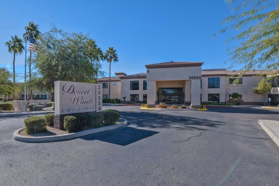Front entrance and driveway of Desert Winds retirement community with a large sign, palm trees, and the building facade.