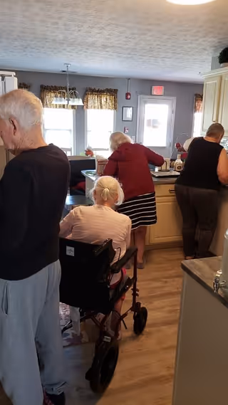 Three elderly women in a kitchen area of a senior living facility. One woman is standing on the left side, another woman in a wheelchair is in the center facing away, and a third woman is standing at the kitchen counter preparing something. The kitchen has light-colored cabinets, a wooden floor, and windows with patterned curtains.