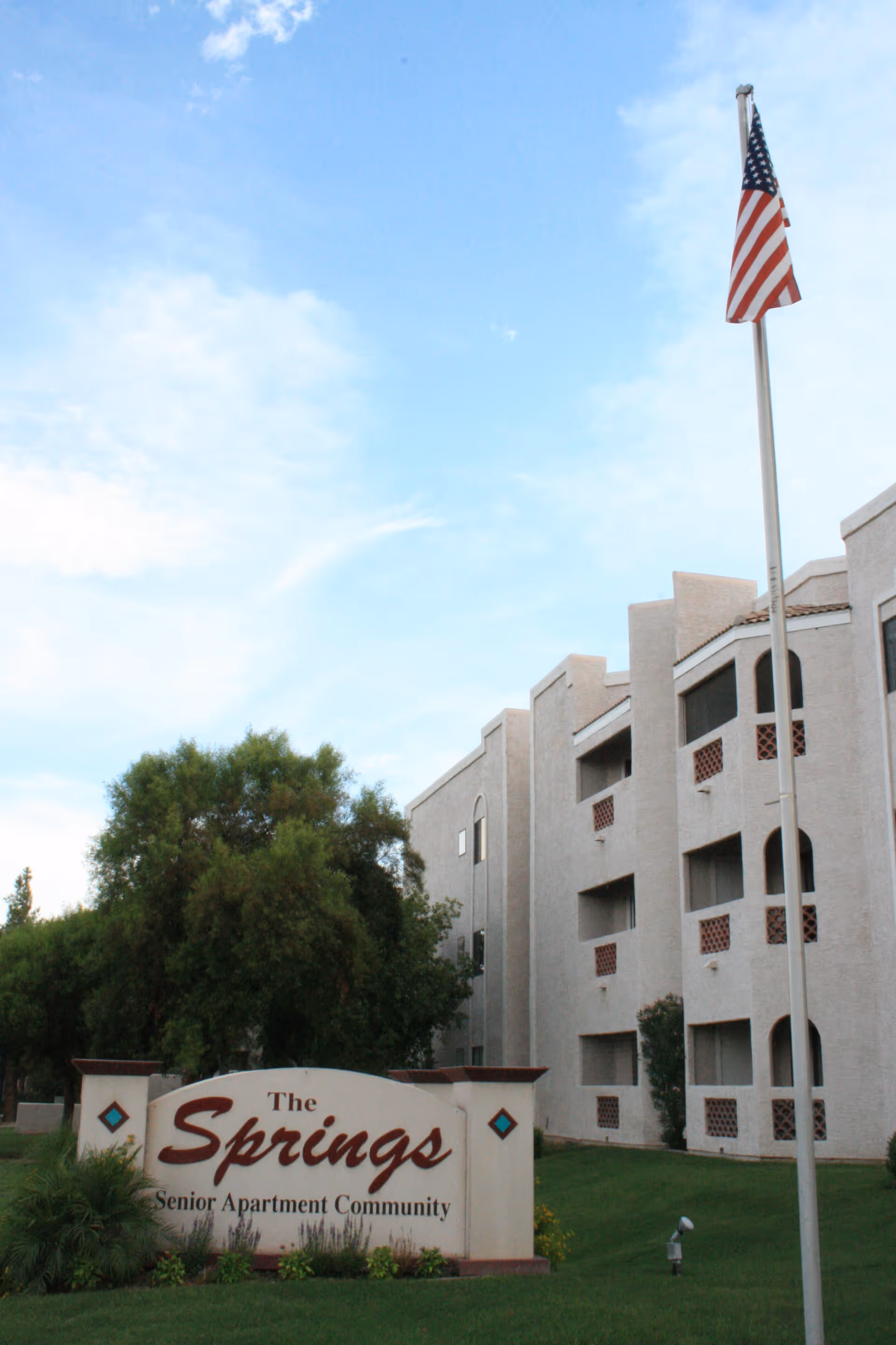 Exterior view of The Springs Senior Apartment Community building with a sign in front, green lawn, trees, and an American flag on a flagpole against a partly cloudy sky.
