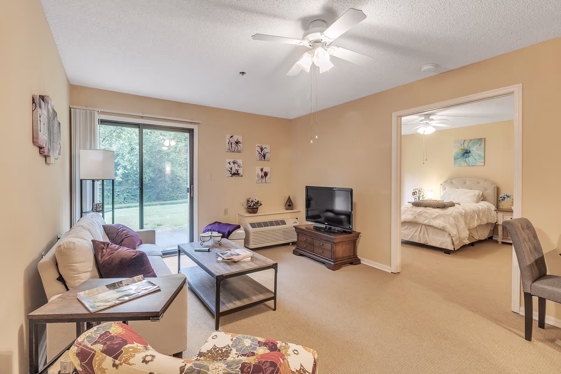 A cozy living room with a beige sofa adorned with purple pillows, a wooden coffee table, a floral patterned armchair, and a TV on a wooden stand. The room has beige walls and carpet, a ceiling fan with lights, and a sliding glass door leading to a green outdoor area. Adjacent to the living room is a bedroom with a neatly made bed, a nightstand, and a ceiling fan.
