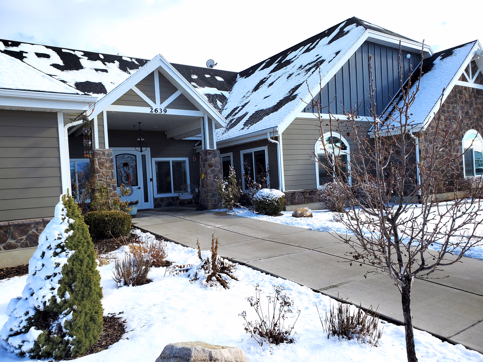 Exterior view of a senior living facility building with snow on the ground and roof. The building has stone and siding walls, a covered entrance with the number 2639 above it, and some leafless trees and shrubs in the snowy landscape.