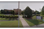 Front exterior of a single-story brick assisted living facility with a central walkway, manicured shrubs, and a lawn sign.