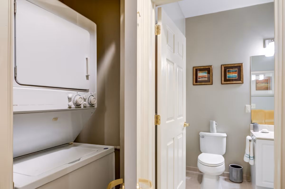 View of a small laundry area with a stacked washer and dryer unit next to a bathroom. The bathroom has a white toilet, a vanity with a sink, a mirror, two framed pictures on the wall, and a small trash can on the floor.